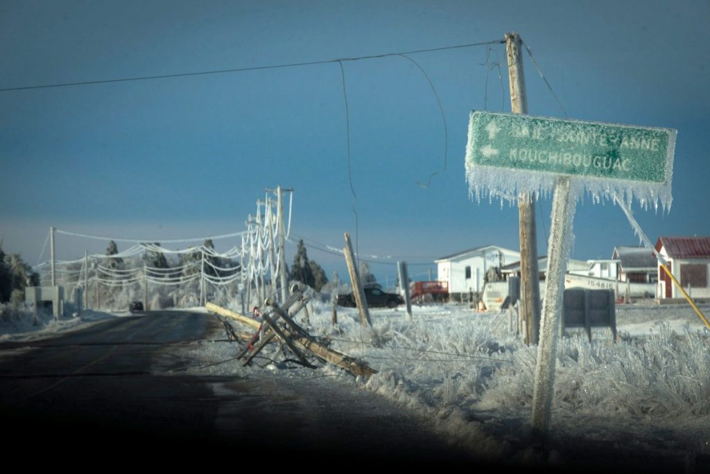 Photograph of a road, lined with utility poles and houses. A road sign pointing toward Kouchibouguac and Baie-Sainte-Anne is visible in the foreground. All of the visible structures are covered in a thick layer of ice. Damaged and felled utility poles are also visible.