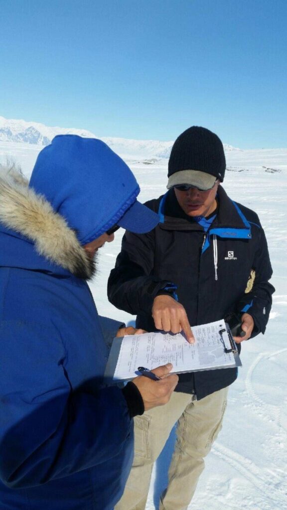 Two community members in winter gear are standing in the foreground of snow-covered foothills and mountains. The landscape appears frigid but under the sun they are comfortably examining a document. One of them is holding a clipboard and pen while the other is pointing to a section the page.
