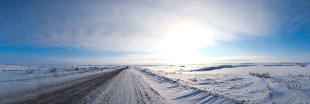 Winter panoramic landscape of an open road, partially covered in snow, in the Inuvialuit Settlement Region. In the horizon, the sun shines through misty clouds casting shadows across the hardened snow.